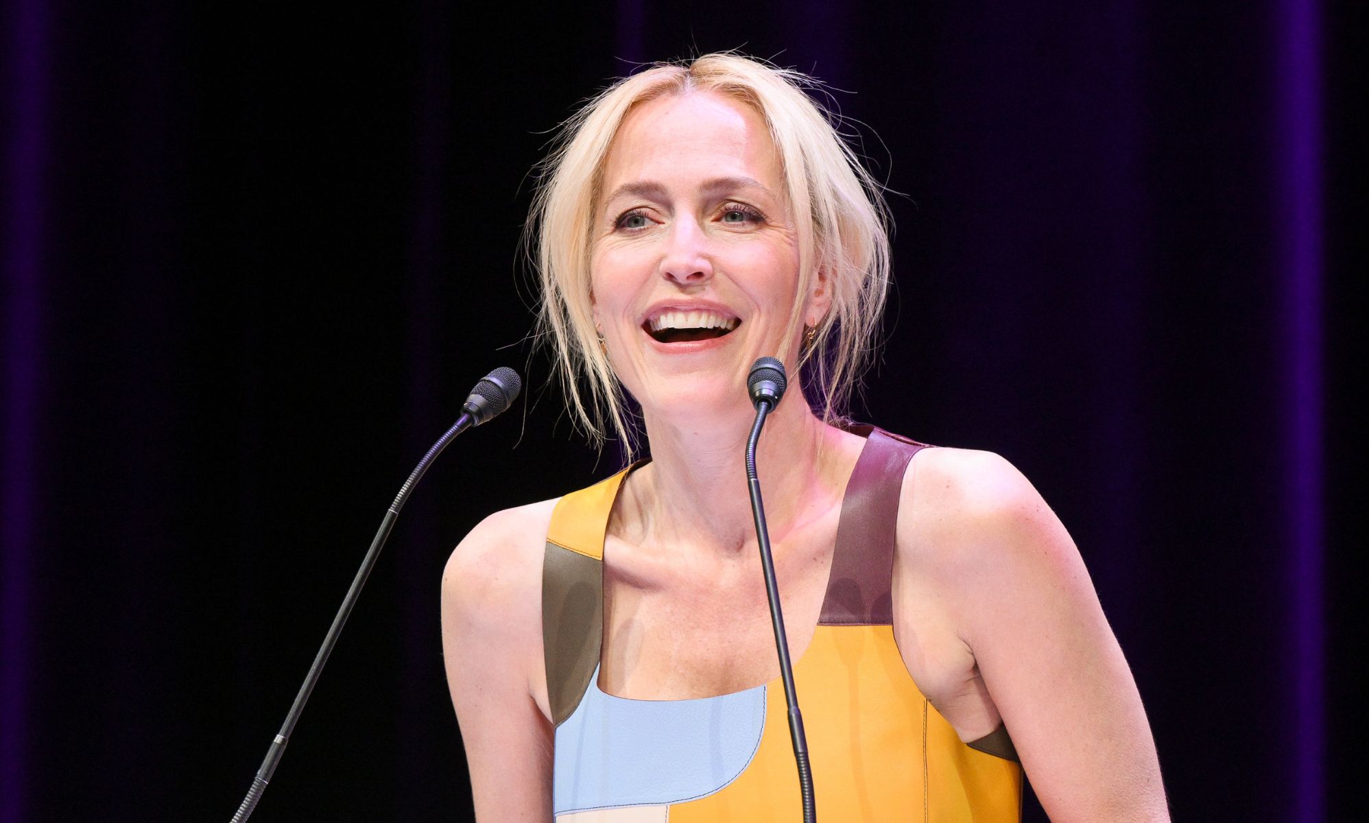 Gillian Anderson in a blue and yellow dress speaking into a microphone.