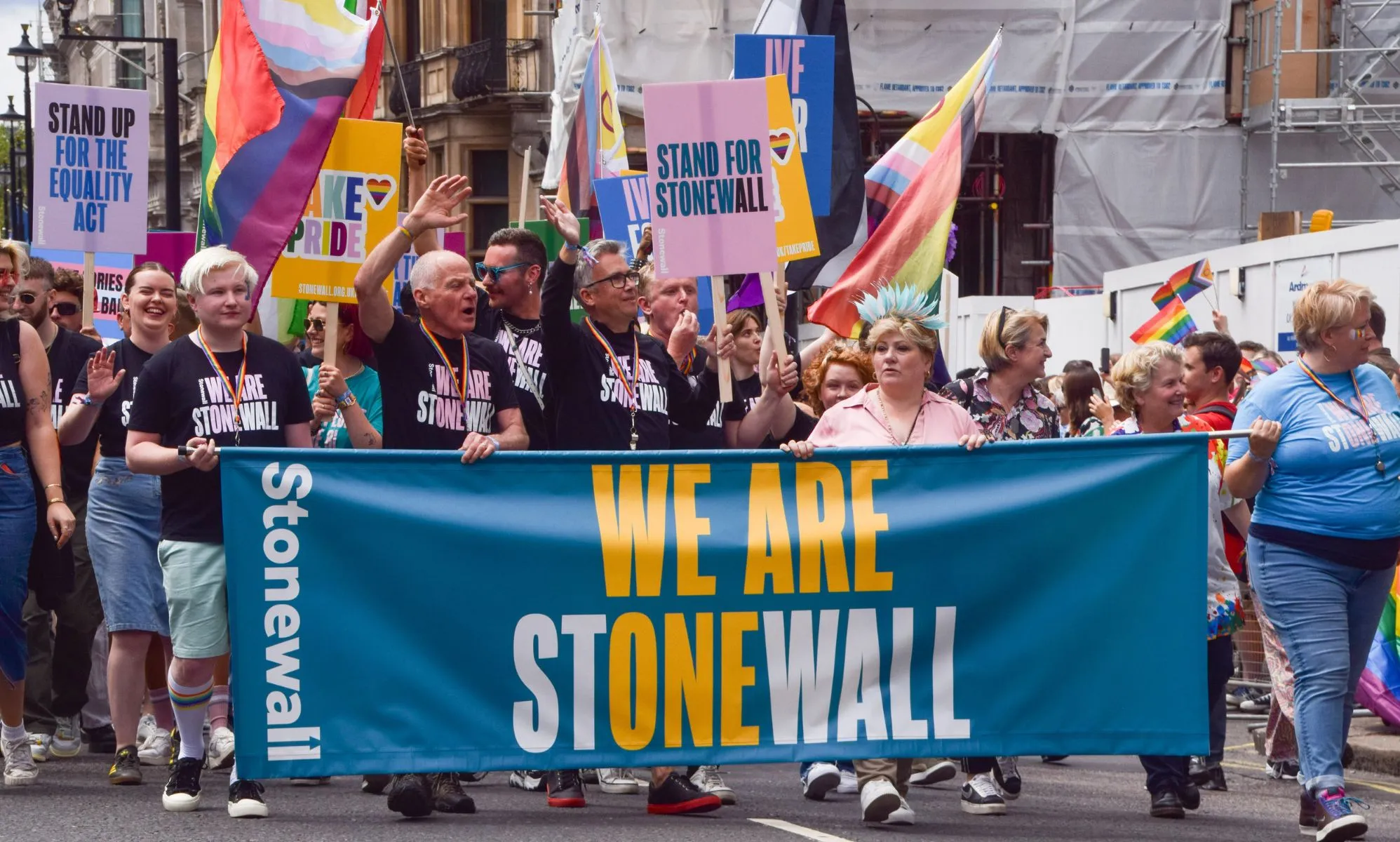 A group of people from LGBTQ+ charity Stonewall stand in a line next to each other as they walk through a Pride march holding up a sign reading