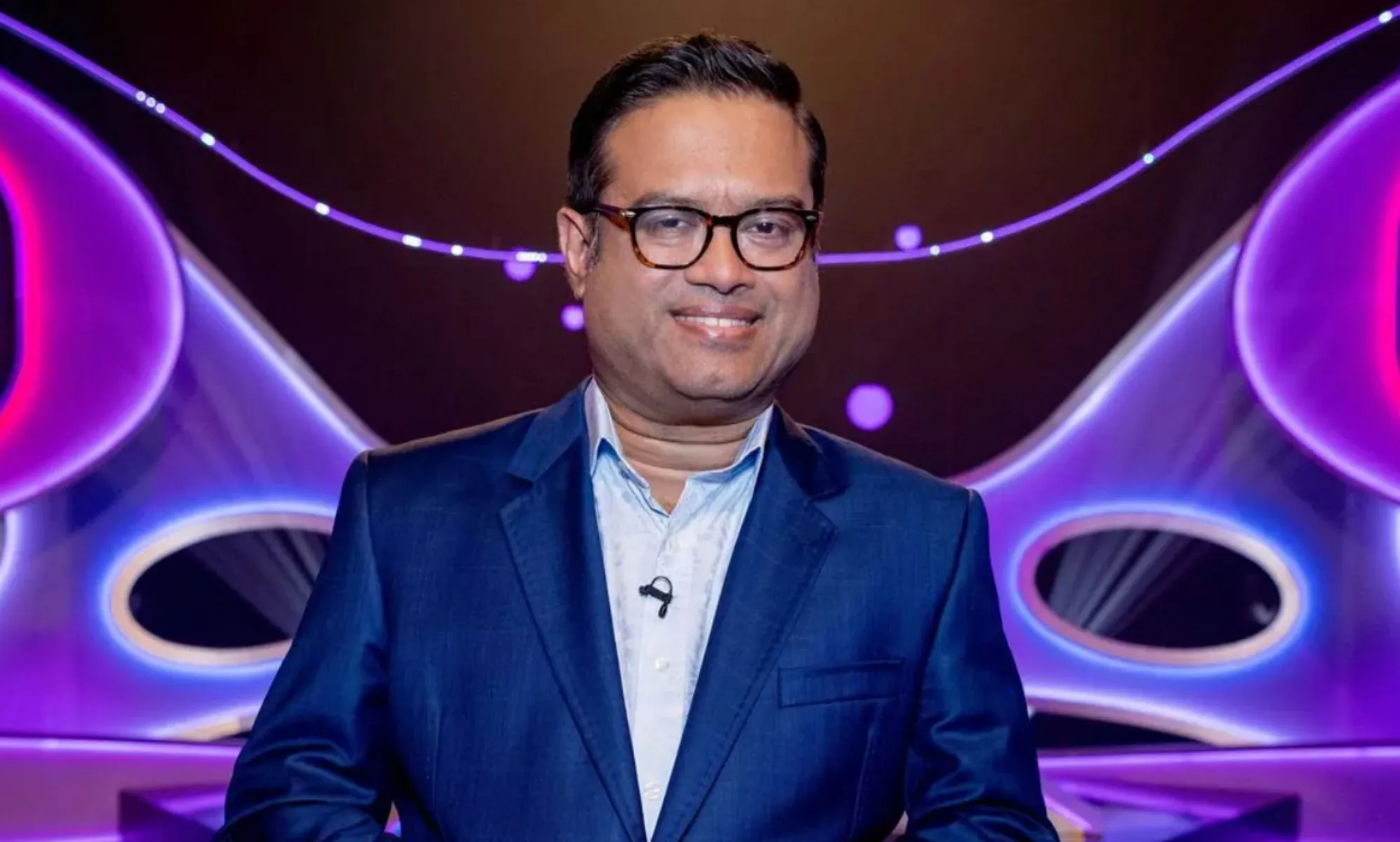 Paul Sinha in a blue suit and shirt standing in front of a blue and purple background.