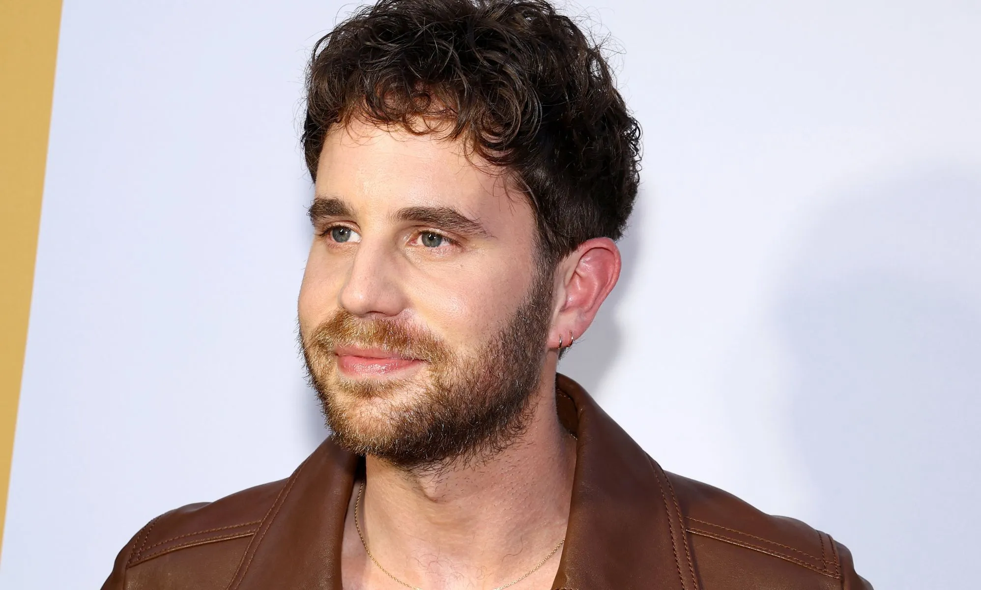Ben Platt, wearing a brown jacket, smiles infront of a white background.