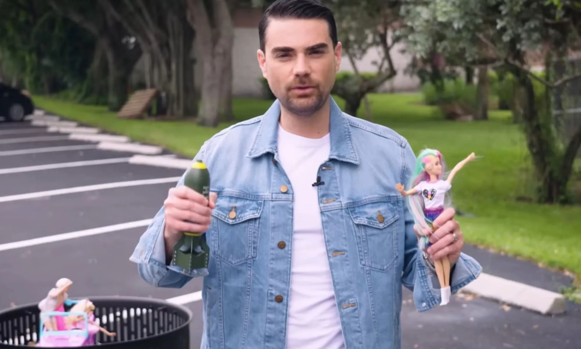 Ben Shapiro holds a Barbie Doll and a miniature replica bomb in the middle of a field.