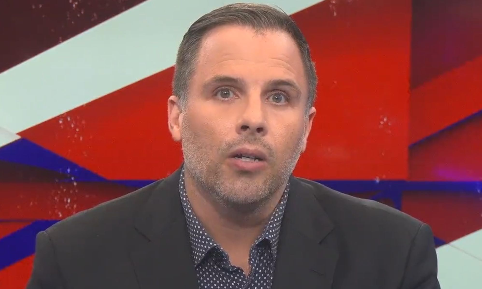 Dan Wootton, on the set of GB News, speaks towards a camera while wearing a suit and open collar shirt.