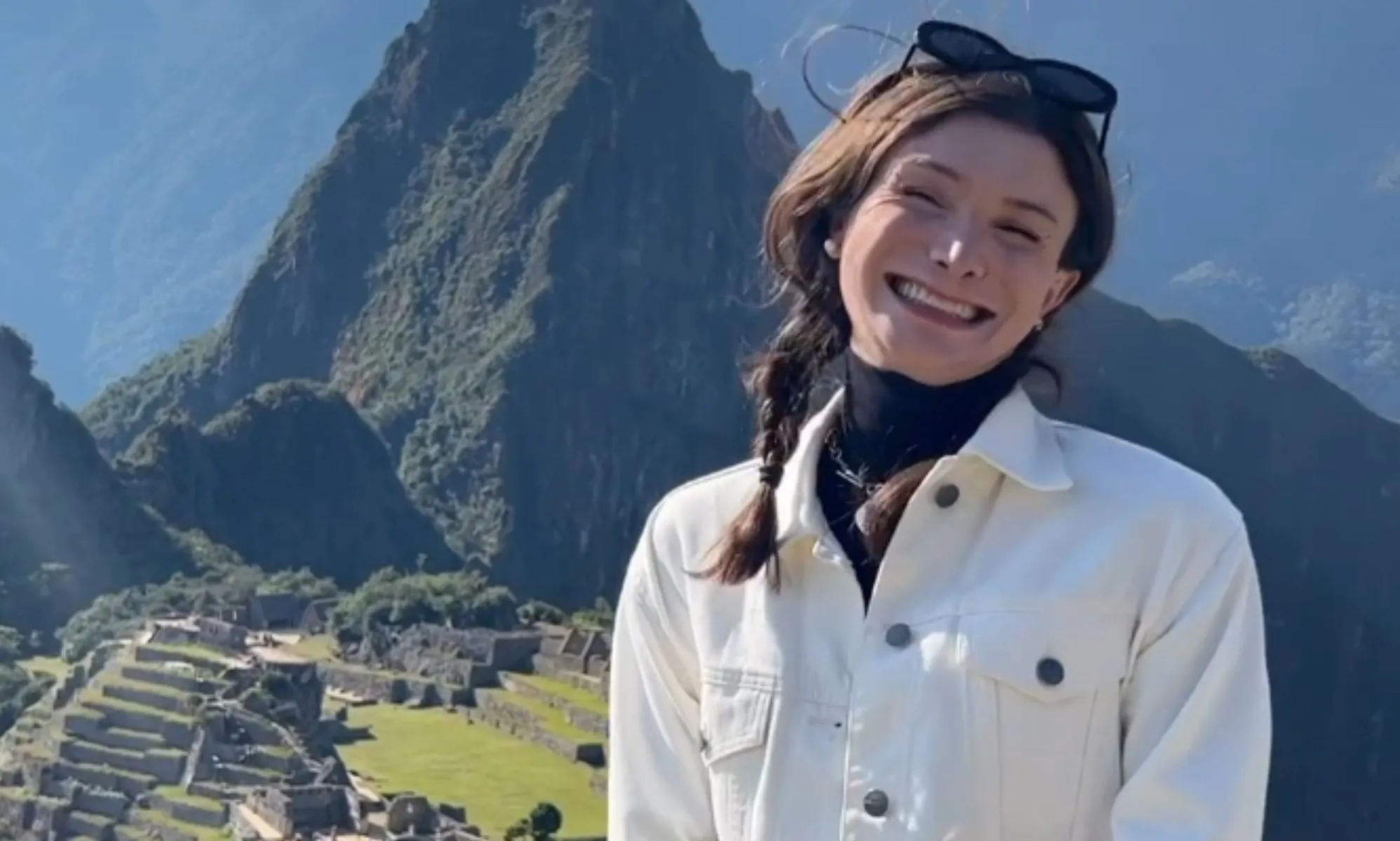 Dylan Mulvaney smiles, while wearing a white jacket, looking over the Peruvian mountains.