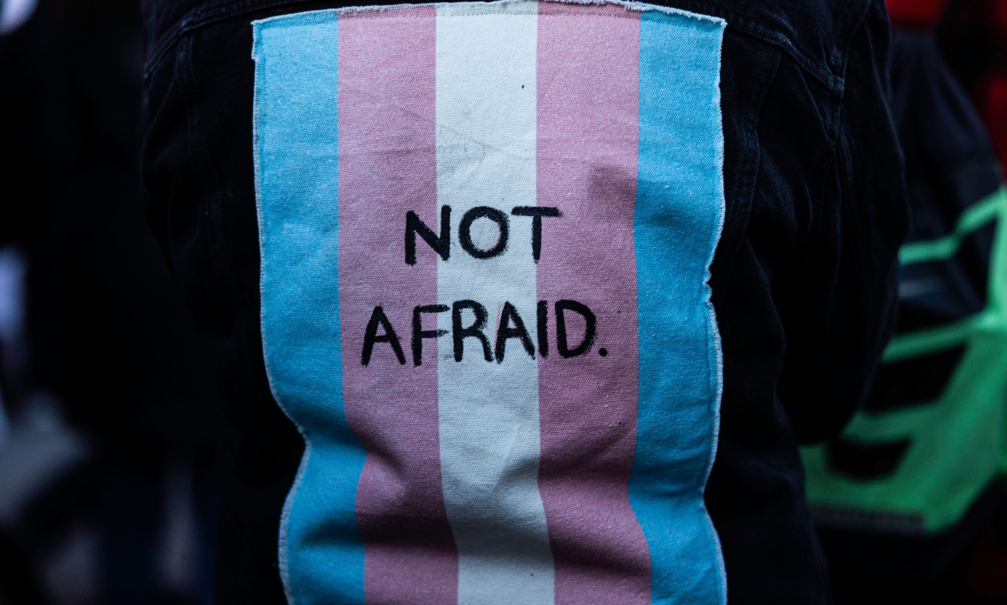 A person wears a jacket with a patch on the back of a trans flag that says