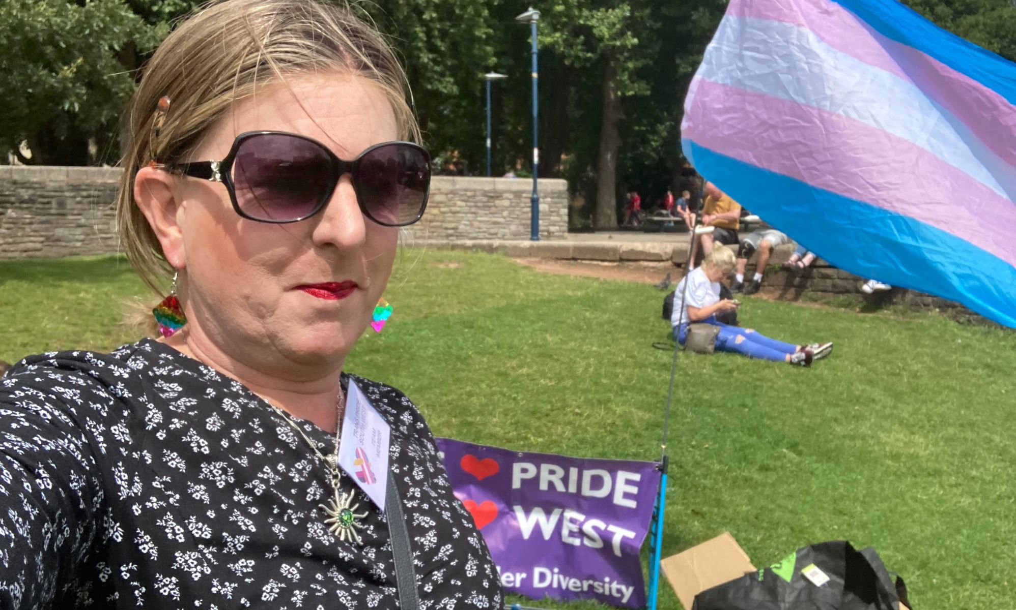 A member of Trans Pride South West taking a selfie in a park next to a trans flag waving in the air.