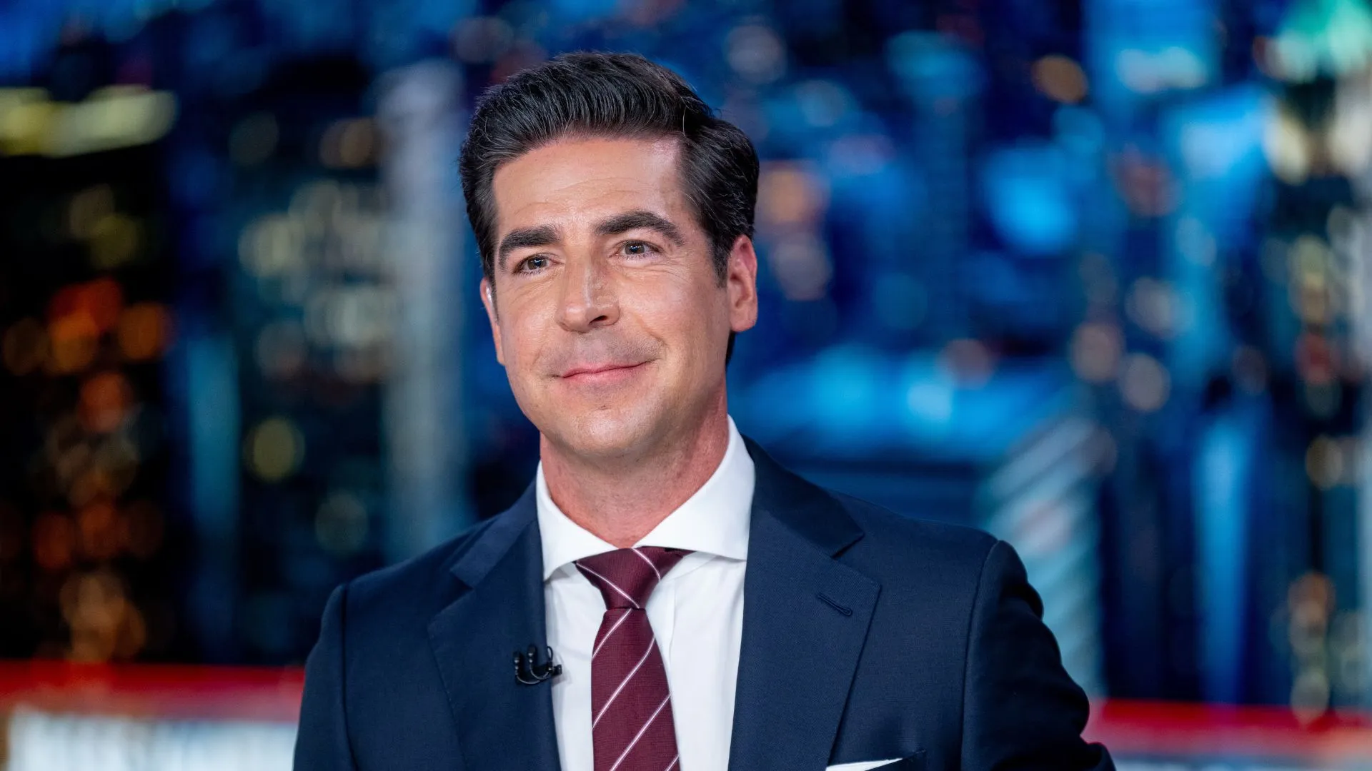 Fox News host Jesse Watters wears a suit and tie and sits at a desk