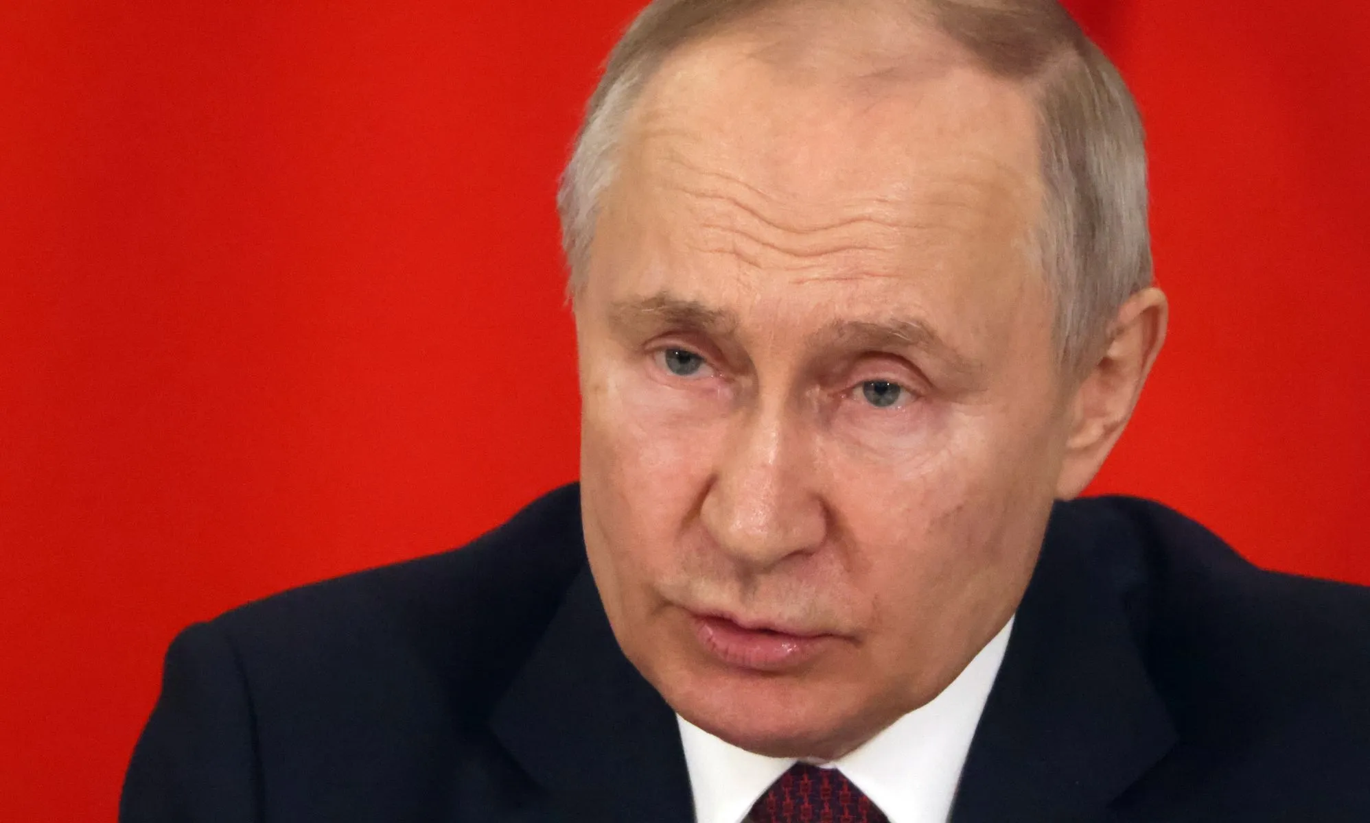 Russia president Vladimir Putin, who has a history of attacking LGBTQ+ people, wears a suit and tie as he sits in front of a red background