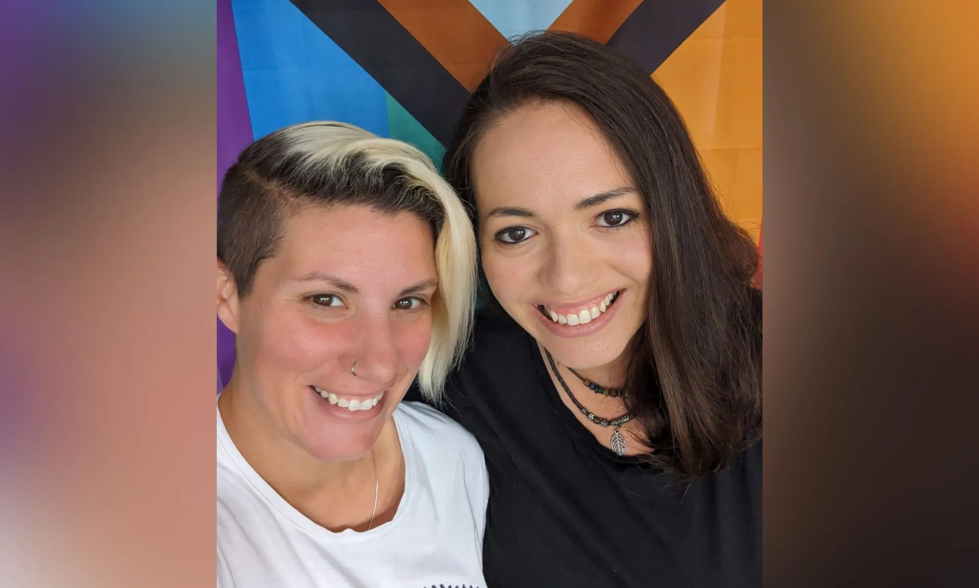LGBTQ+ foster carers Nat and Terri stand side-by-side as they smile for the camera in front of a Progressive Pride flag