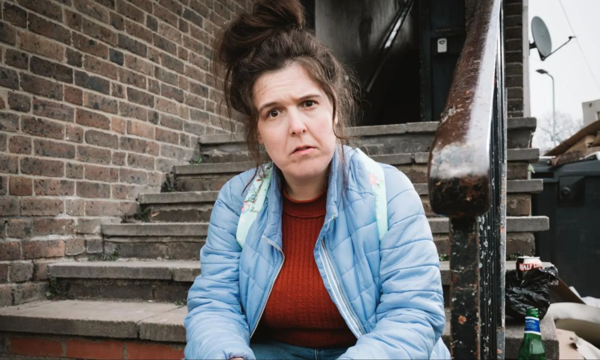 Comedian Rosie Jones as Emily in new sitcom Disability Benefits. She is staring with a questioning glance at the camera, while sitting on some steps in a back alley.