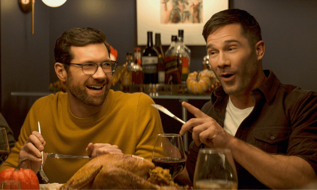 Billy Eichner y Luke Macfarlane en una mesa, riéndose con los cubiertos levantados a medio gesto, en Bros.