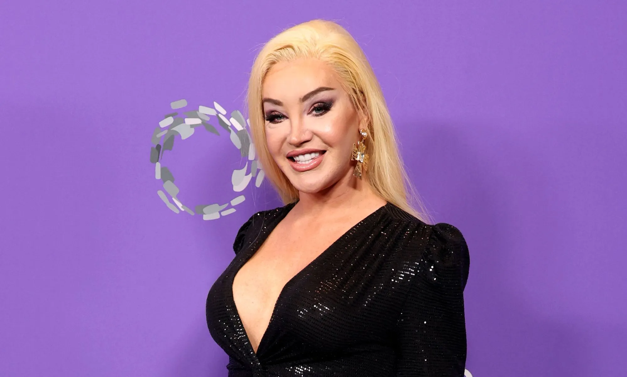 Kylie Sonique Love wears a black dress to an LGBT Gala in 2023. She is smiling at the camera and standing against a purple background.