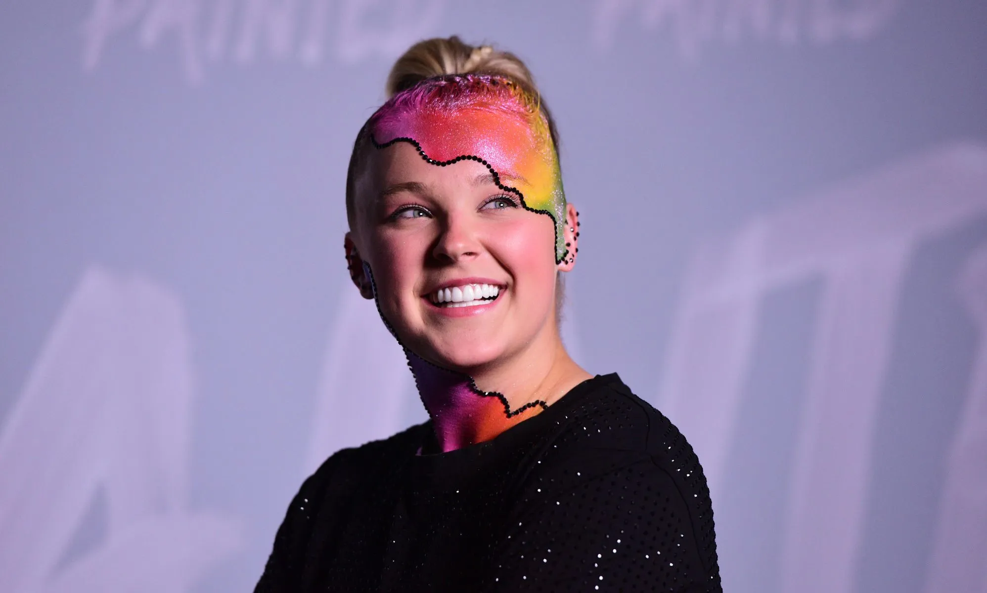 JoJo Siwa wears a black top as she poses with rainbow makeup on her head and neck