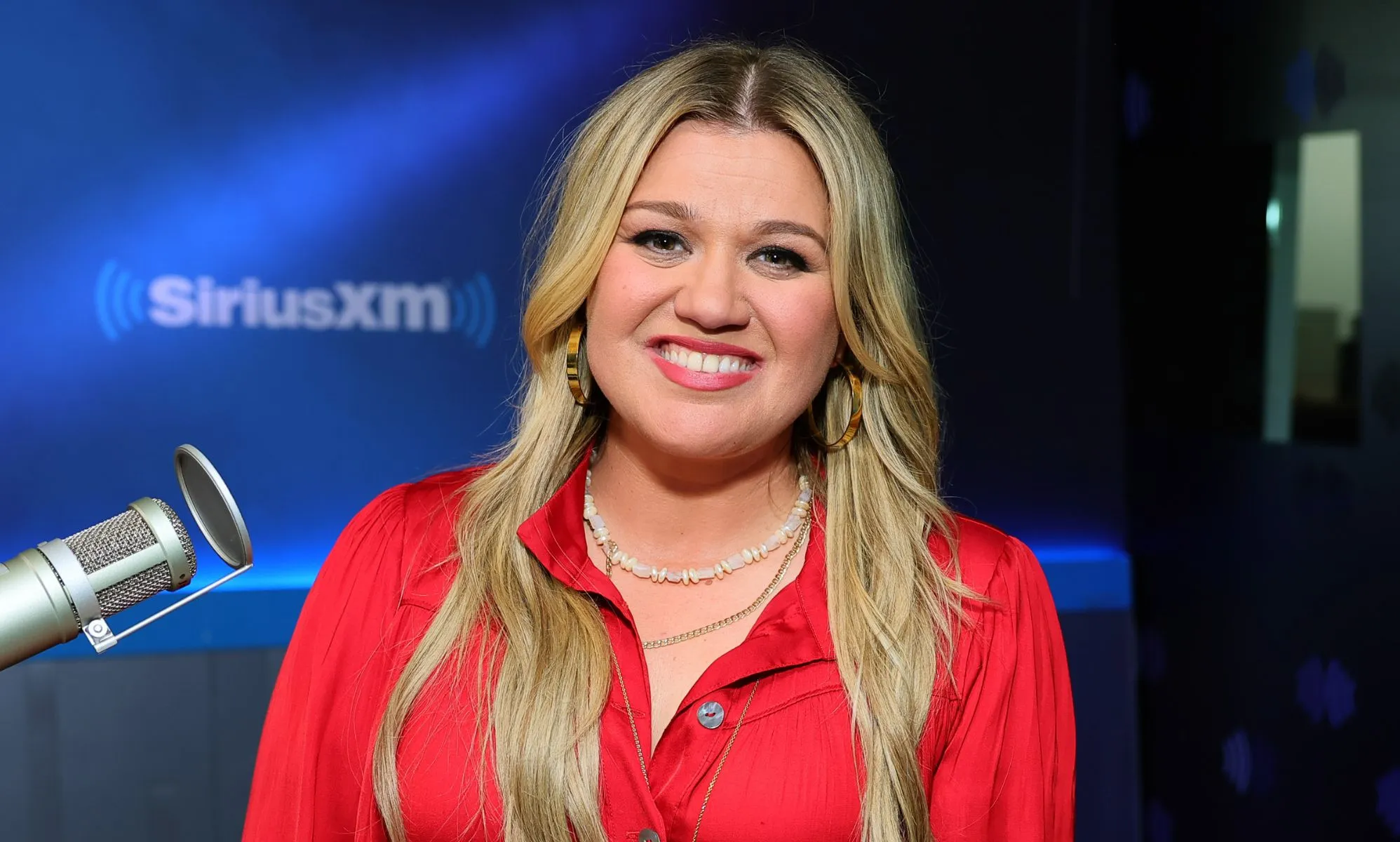 Kelly Clarkson smiling during a radio appearance. She is wearing a red shirt and two necklaces.