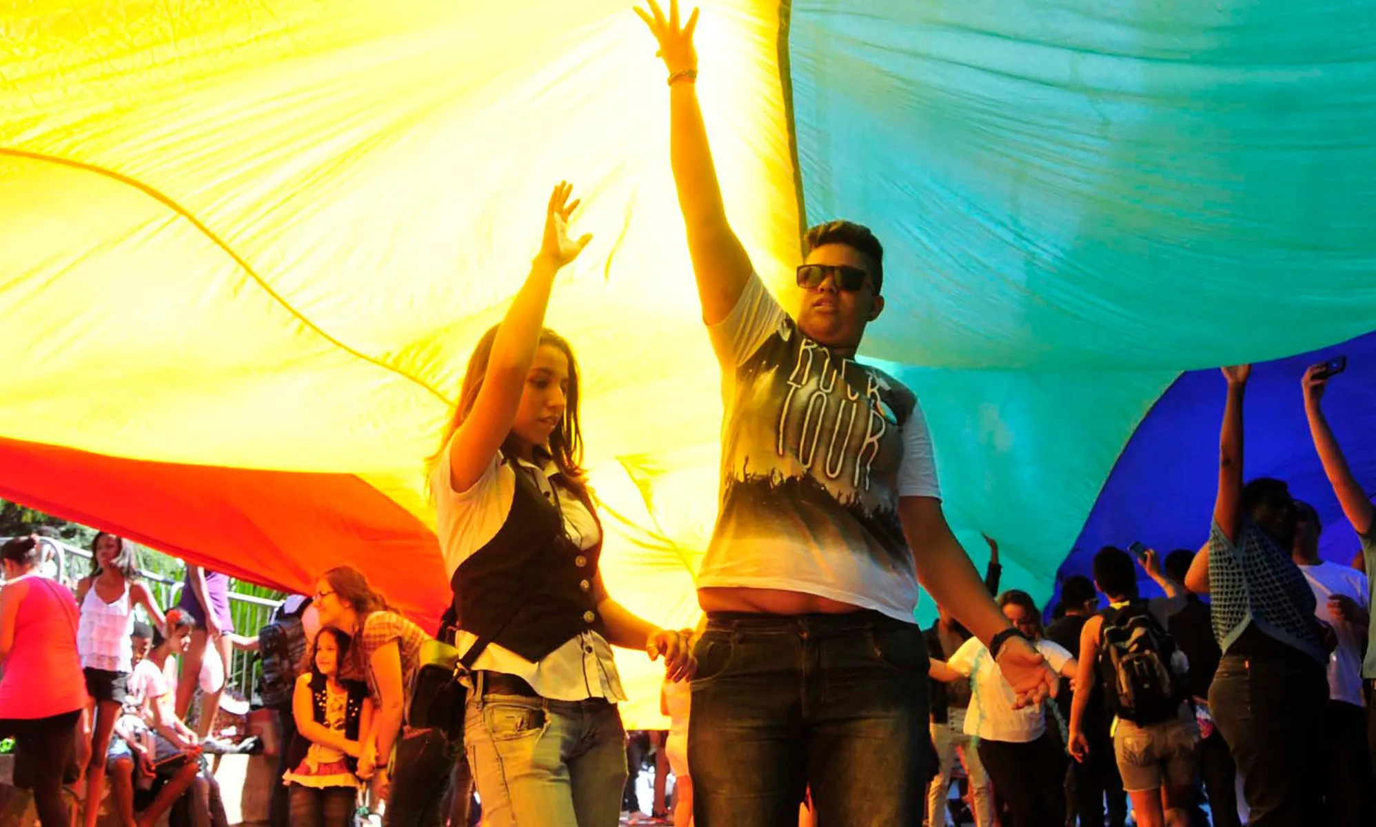 Crowds celebrate during a Brazil Pride march under a giant LGBTQ+ flag.
