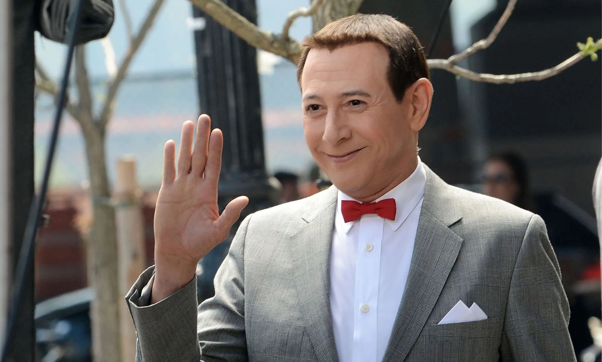 Pee-wee Herman actor Paul Reubens wearing a grey suit, white shirt and red bow tie. He is smiling and waving off camera.