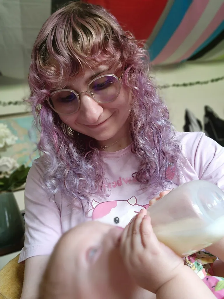 Aquí se muestra a Naomi alimentando a su hijo con leche extraída.  Tiene el pelo rosado, lleva una camiseta rosa y mira con amor a su hijo.