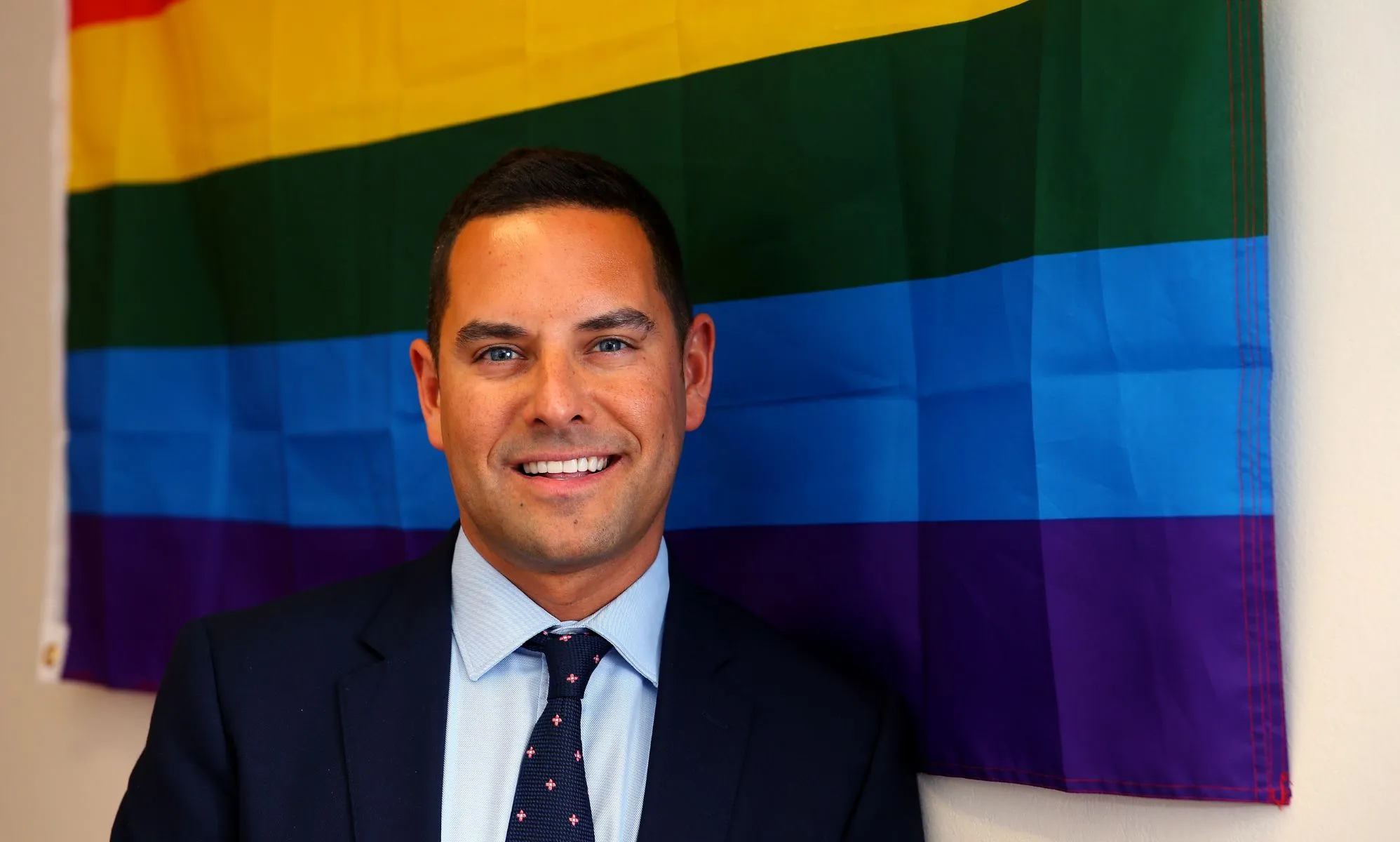 Openly gay Australian MP Alex Greenwich, who has received homophobic hate online, wears a suit and tie as he stands in front of a rainbow LGBTQ+ Pride flag