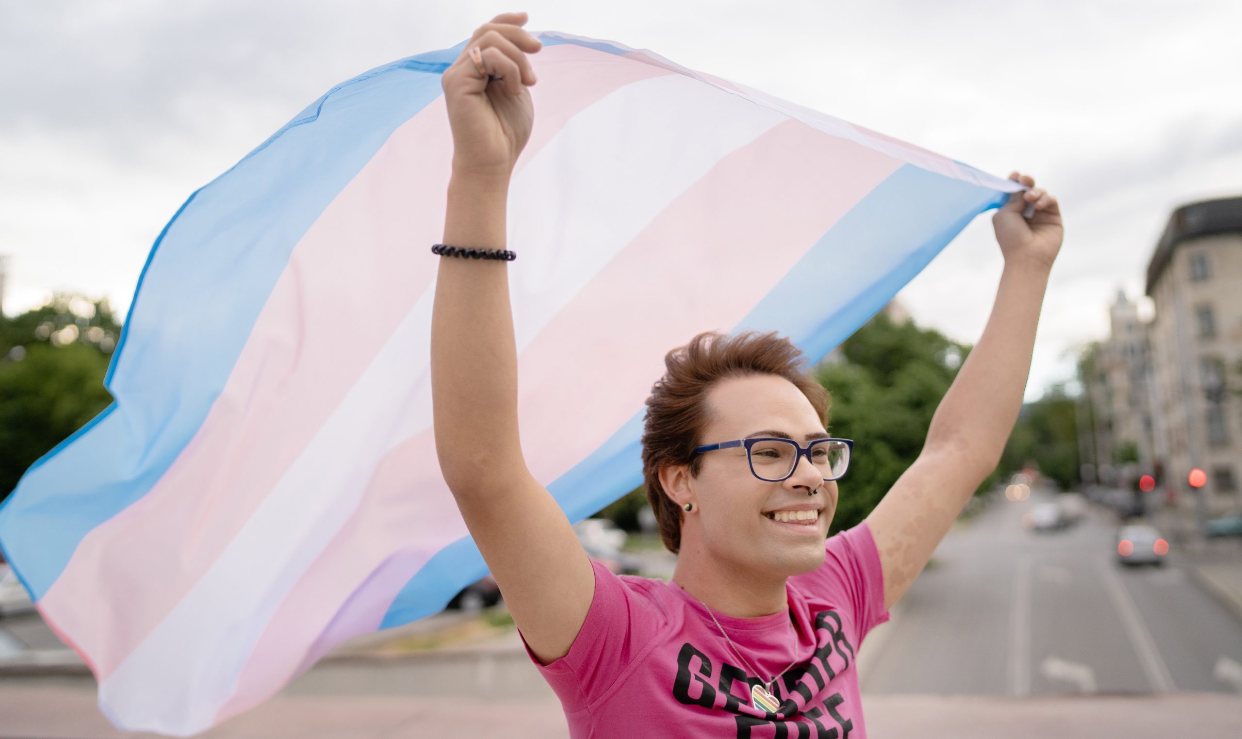 Person wearing a pink shirt and smiling, waving a transgender Pride flag behind them