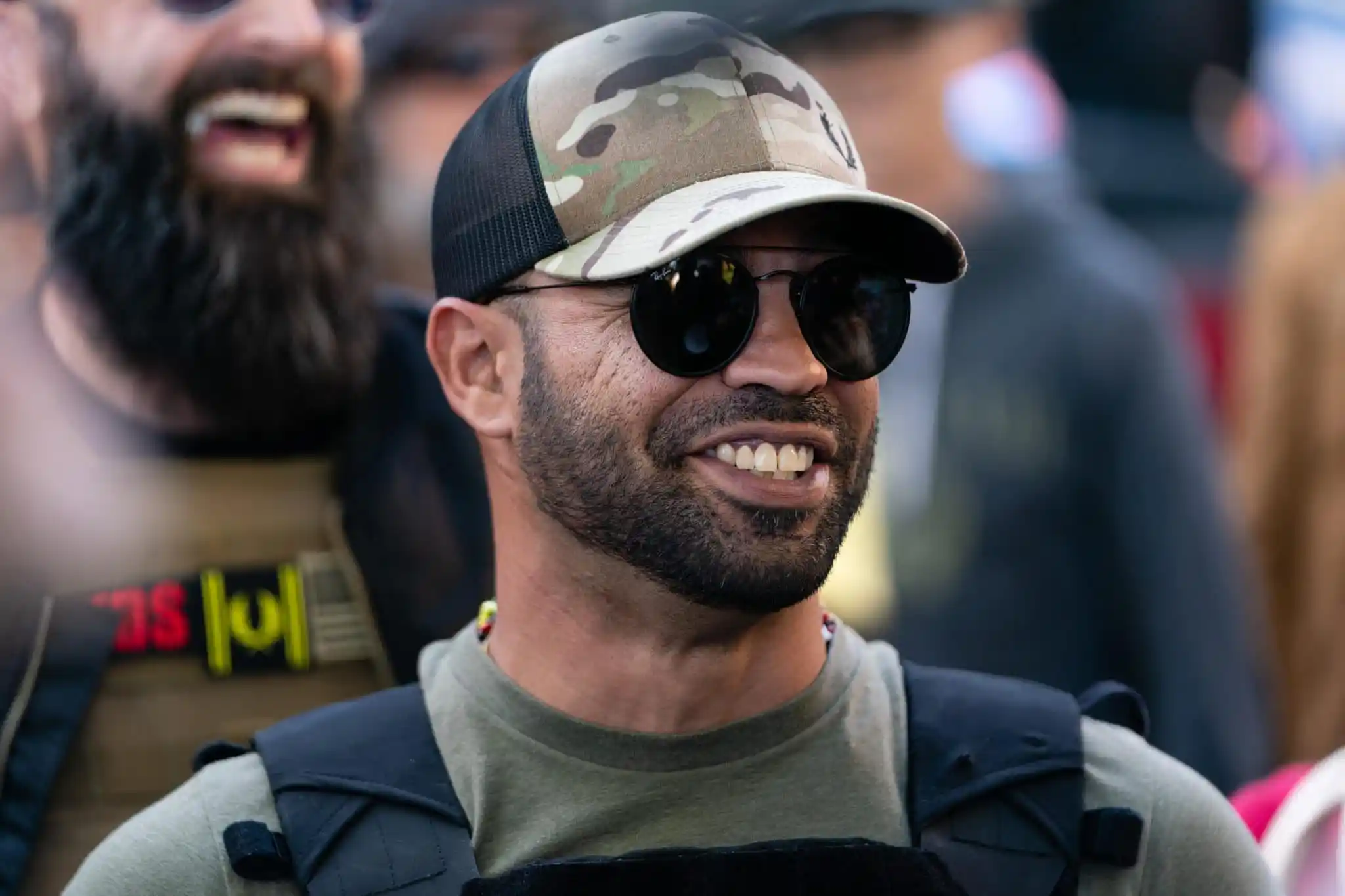 Proud Boy leader Enrique Tarrio smile while wearing a dark green t-shirt and cap