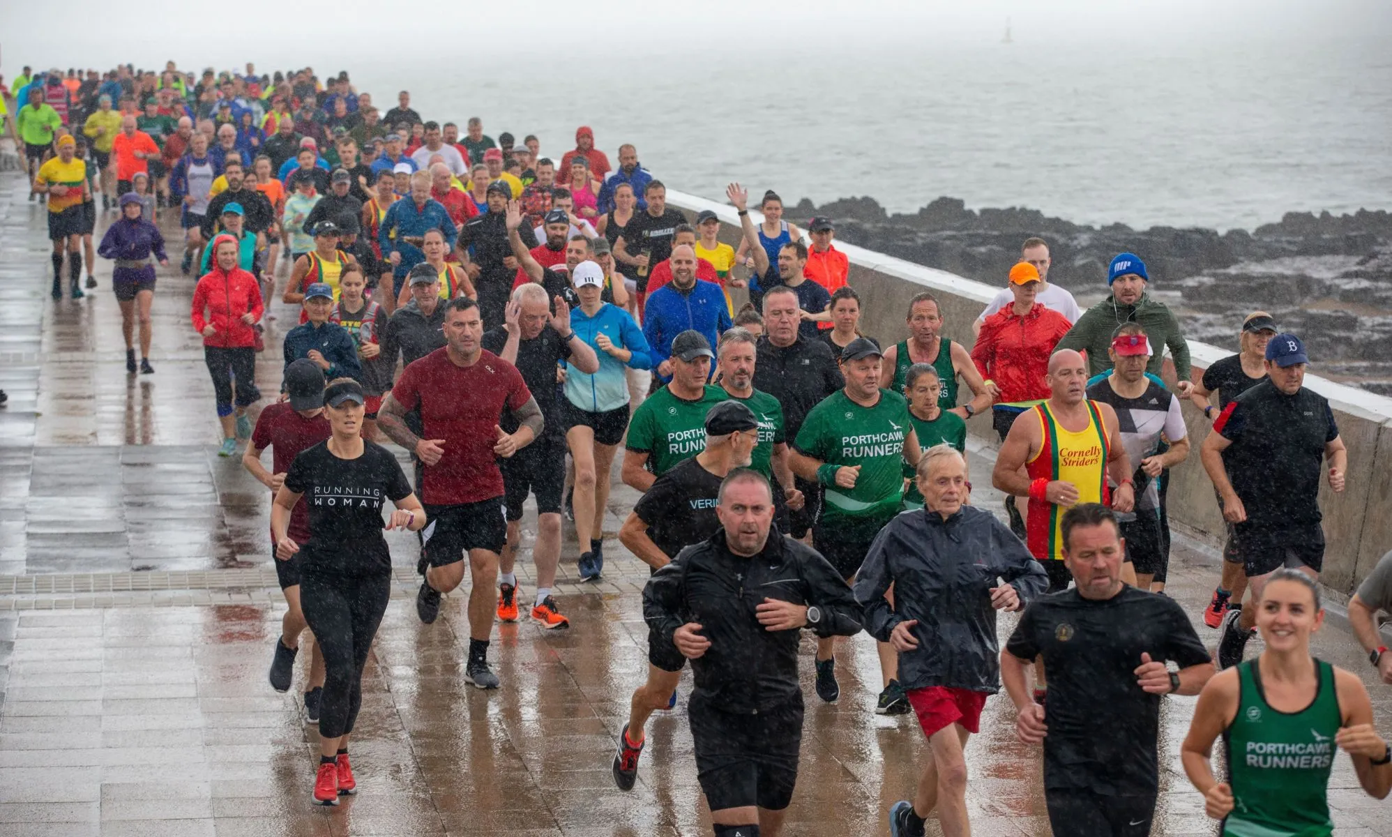 People taking part in a Parkrun in Wales. The image shows many people running on a wet, rainy day by the sea.