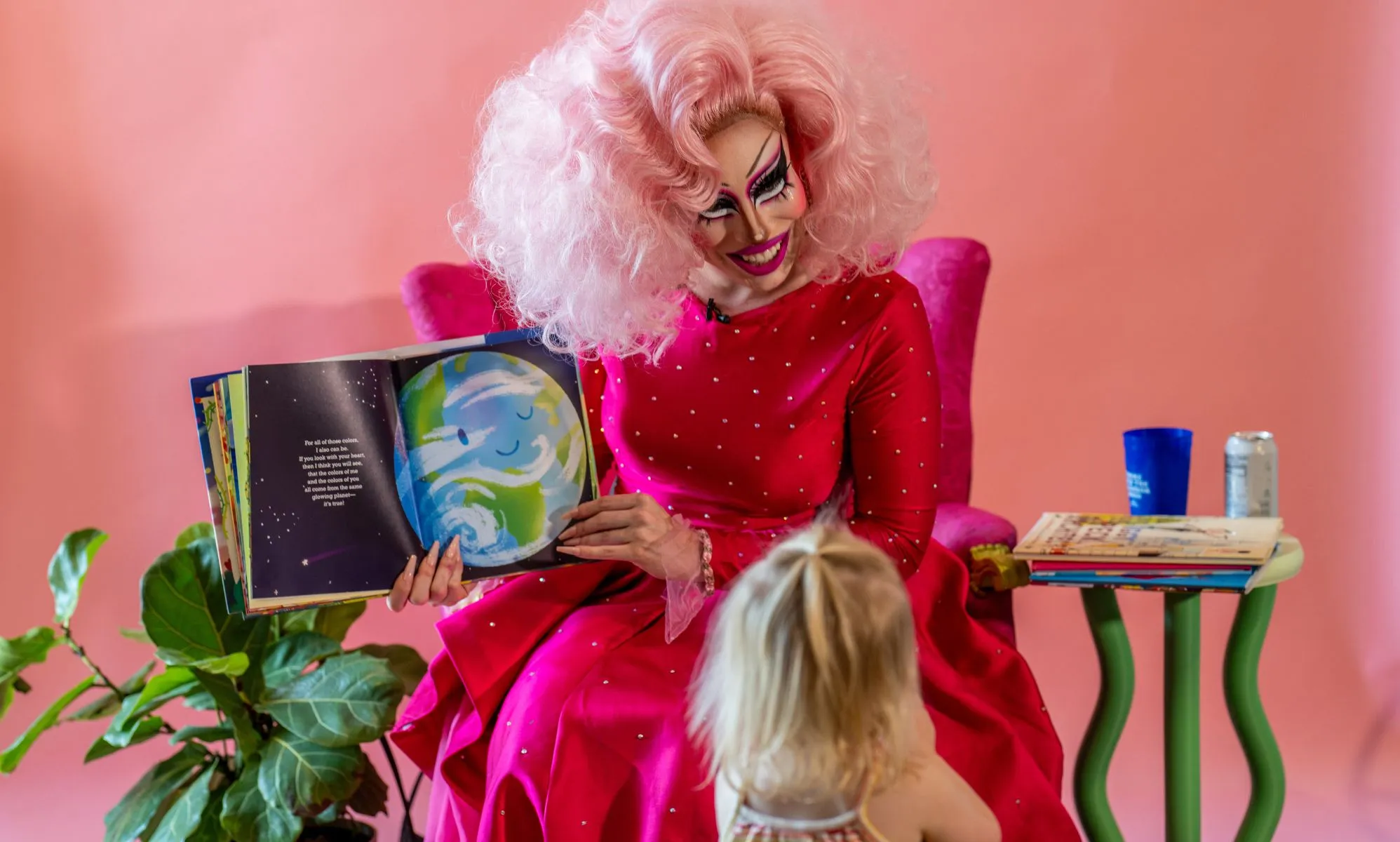 A drag queen, dressed all in pink, holds up a book while interacting with kids during a story time reading event