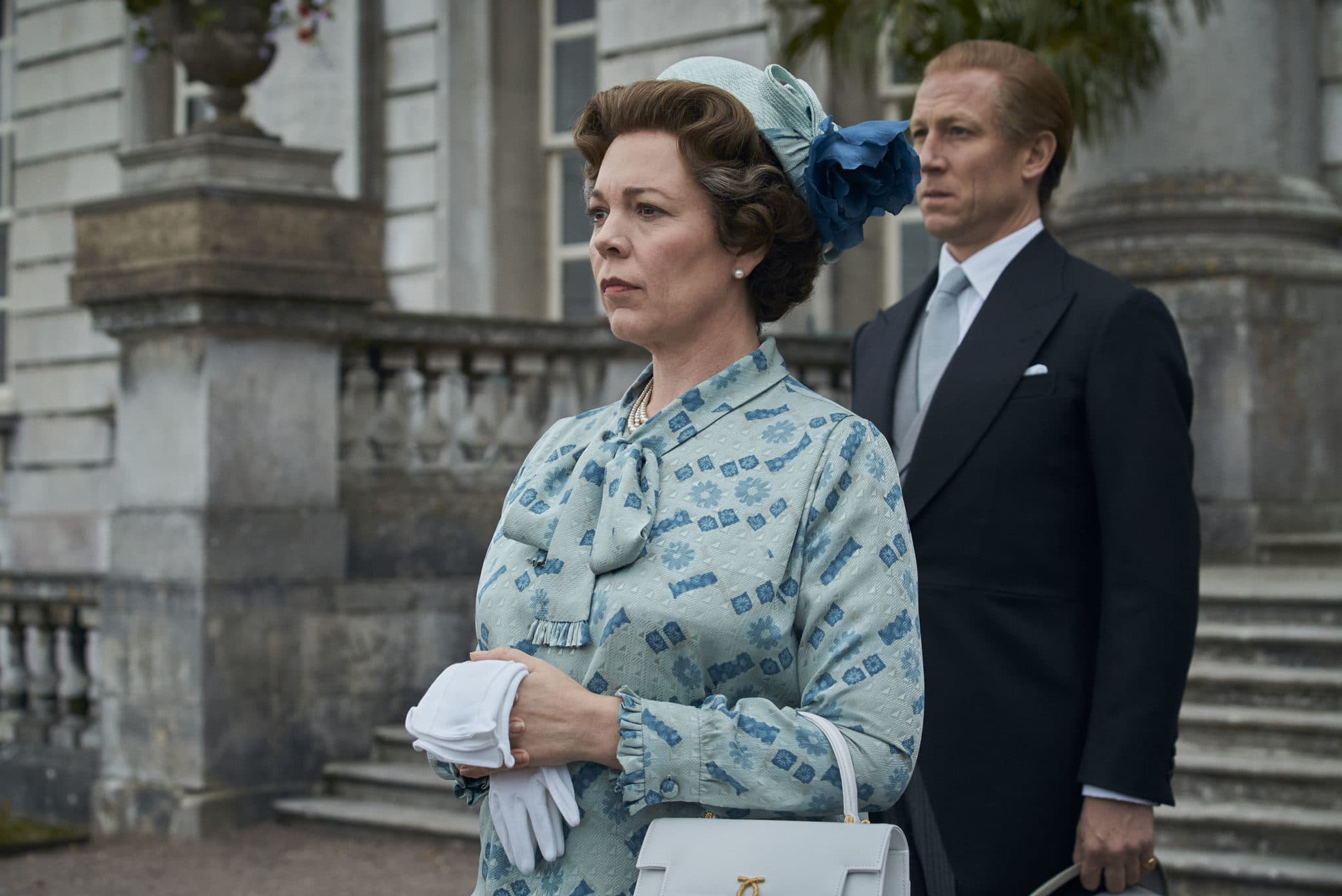 Olivia Colman as the Queen standing on the steps of the palace with Prince Philip behind her, in a duck-egg blue pussy-bow and pillbox hat