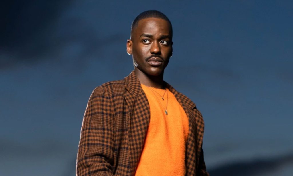 Ncuti Gatwa in a promotional photo for Doctor Who. He is wearing a brown and black chequered jacket and orange top with a necklace. He is looking off camera. The sky is behind him.