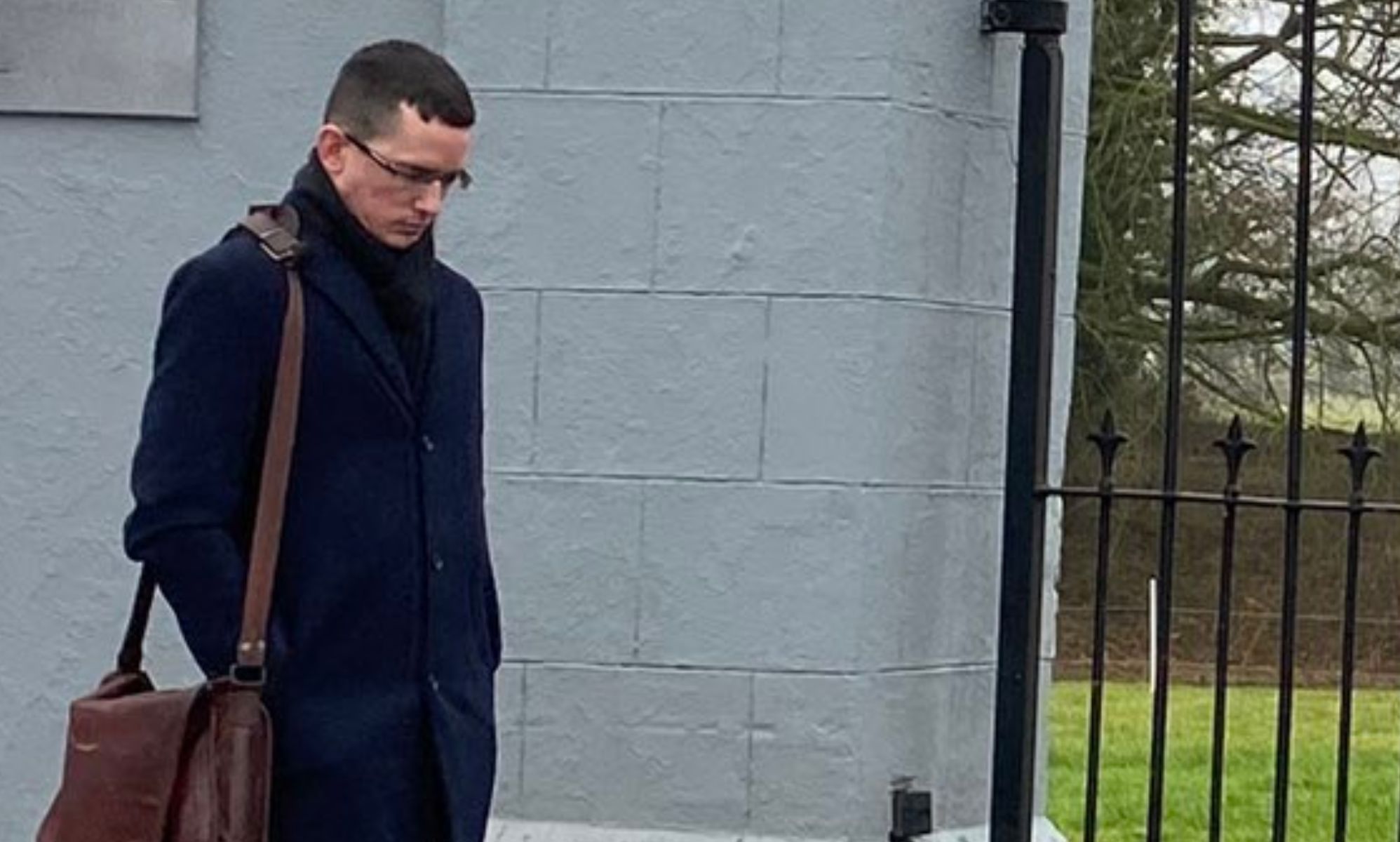 Enoch Burke stands beside a grey school gate, looking down on the floor.