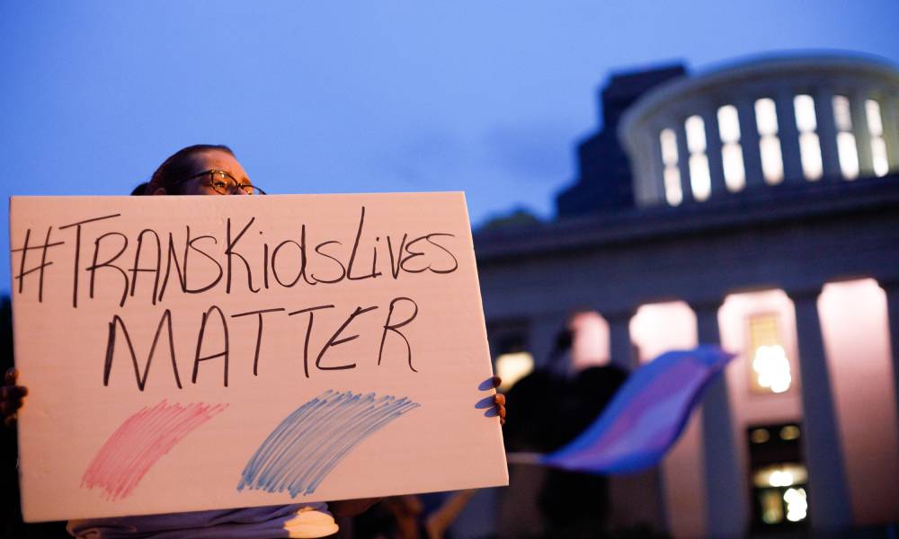 Un defensor de los derechos de las personas transgénero sostiene un cartel que dice '#TransKidsLives Matter' con colores rosa y azul debajo mientras se encuentran afuera del Capitolio de Ohio durante una manifestación contra un proyecto de ley que prohíbe los deportes trans.