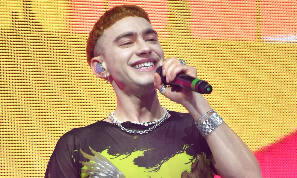 Olly Alexander wears a yellow and black shirt as he sings into a microphone while on stage