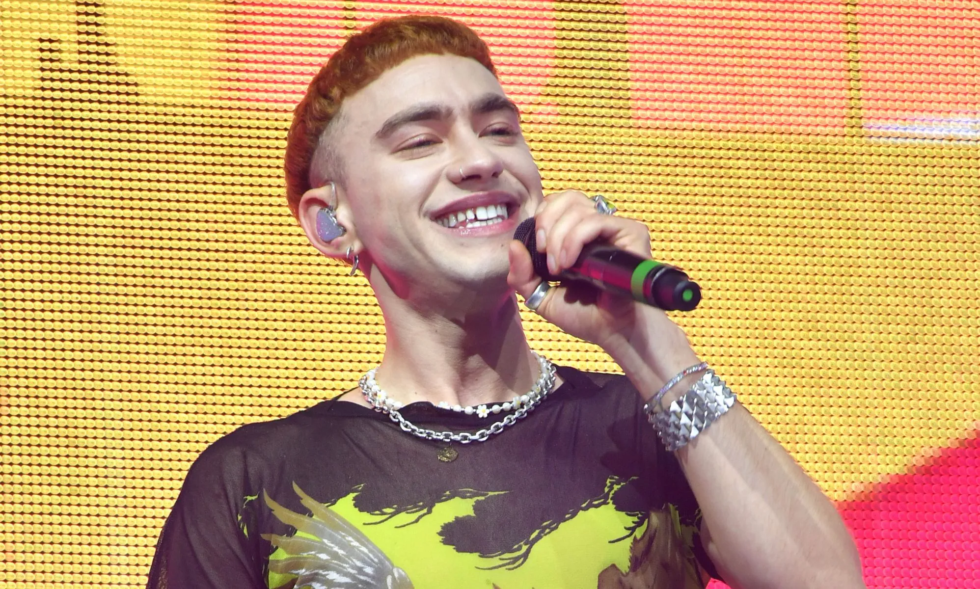 Olly Alexander wears a yellow and black shirt as he sings into a microphone while on stage