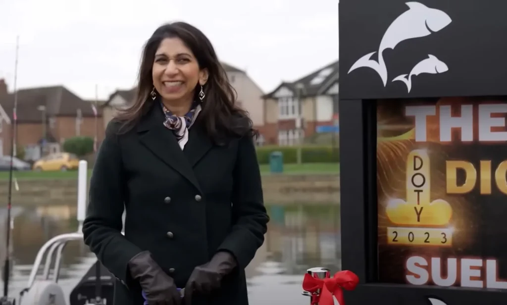 Former home secretary Suella Braverman, wearing a black coat, smiles awkwardly, as she