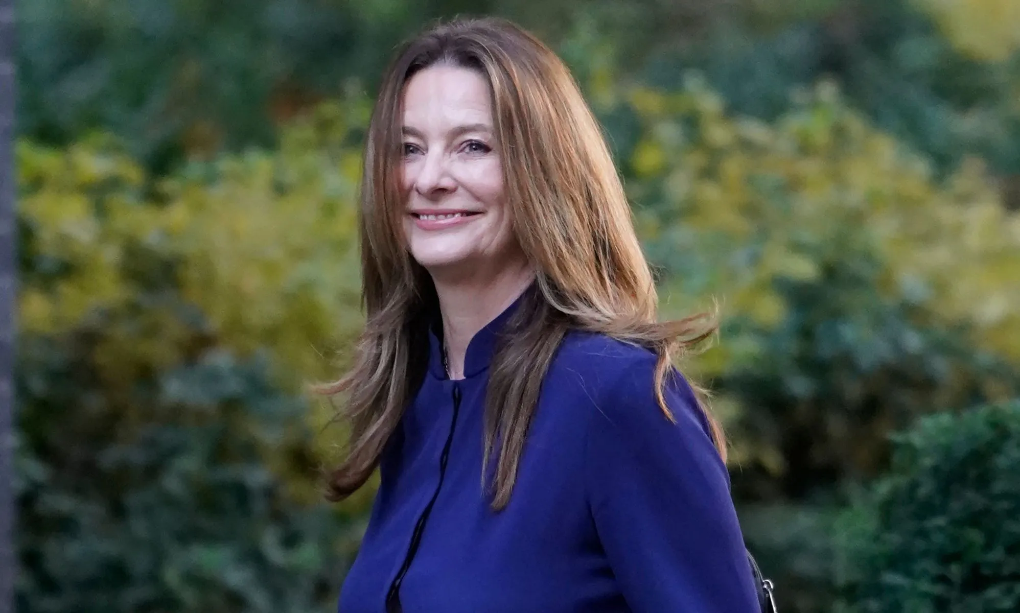Gilian Keegan, in a navy blue dress, walks towards number 10 downing street by nearby foliage, which is blurred in the background.
