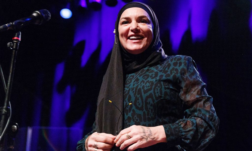 Sinéad O'Connor lleva un vestido negro y verde mientras sonríe cerca de un micrófono.  Ella se encuentra entre las estrellas y aliados LGBTQ+ que murieron en 2023.