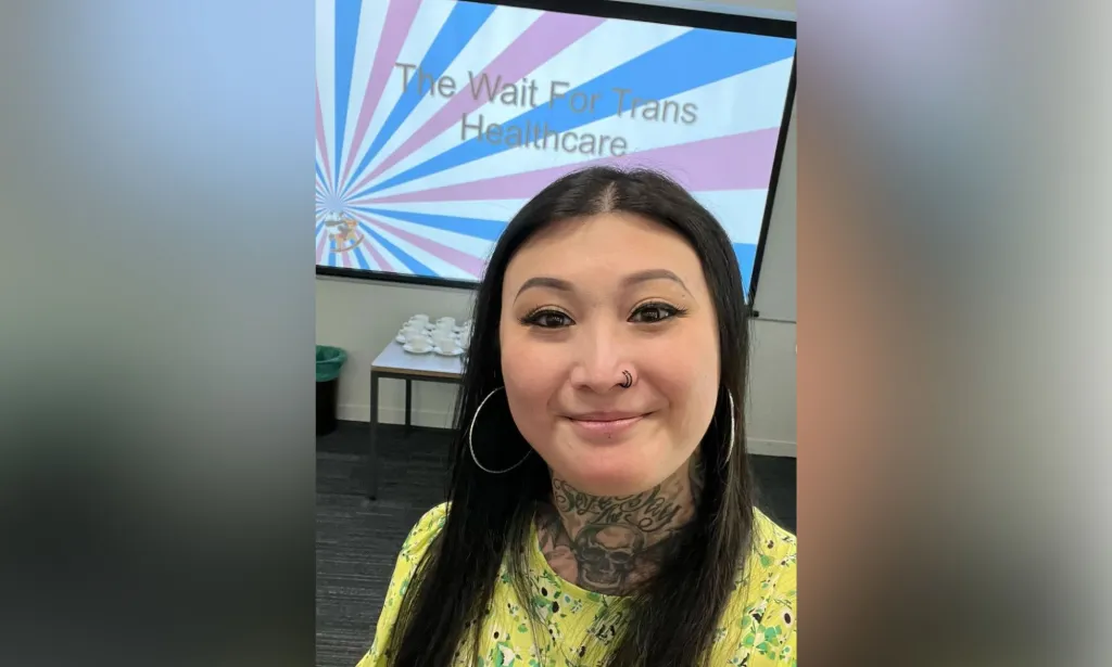 Eva Echo sonríe a la cámara mientras usa una blusa estampada amarilla y verde con una pantalla al fondo que dice 'The Wait For Trans Healthcare' con los colores de la bandera del Orgullo trans.
