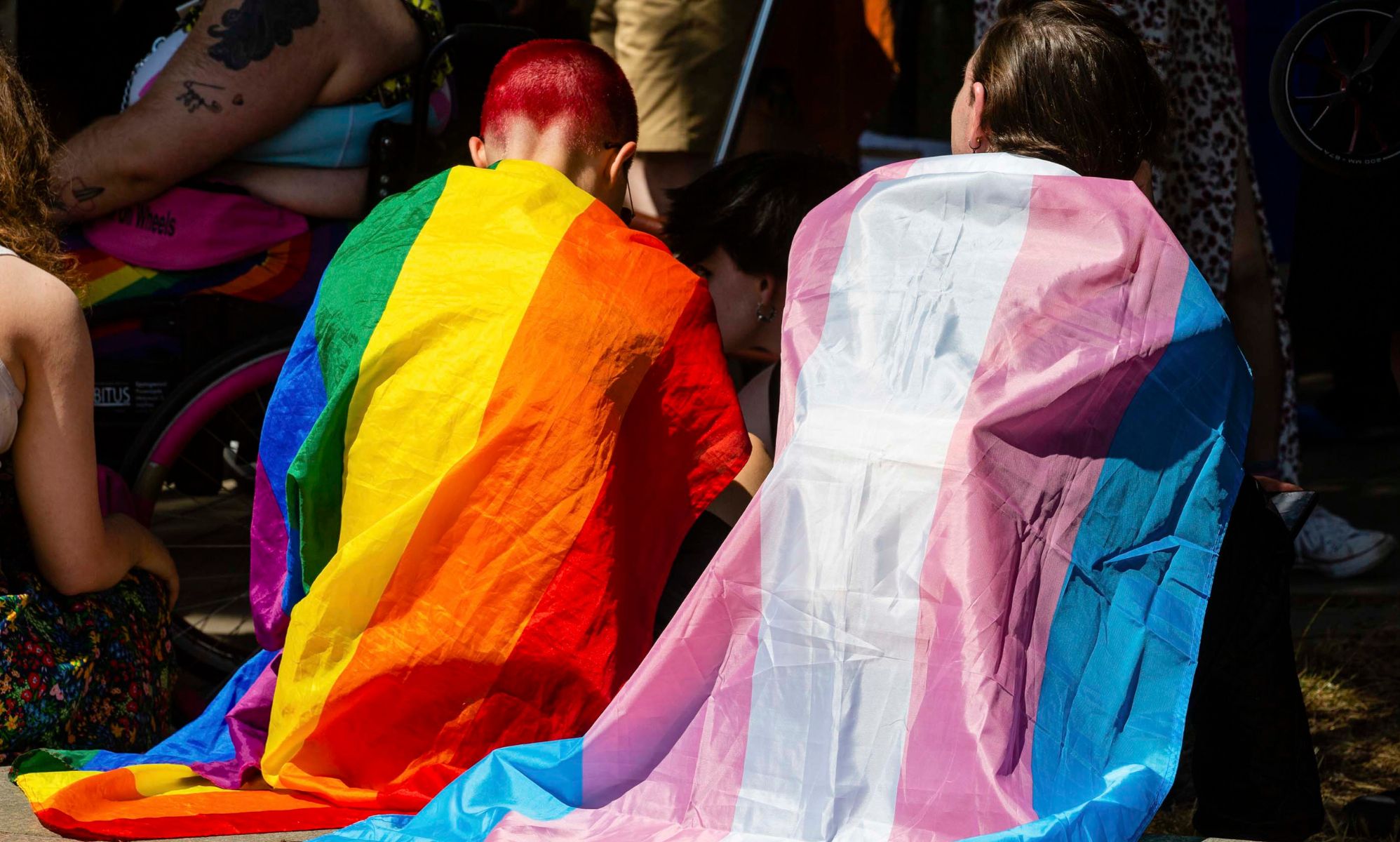 Two people sit together wearing a trans flag and a Pride flag as capes.