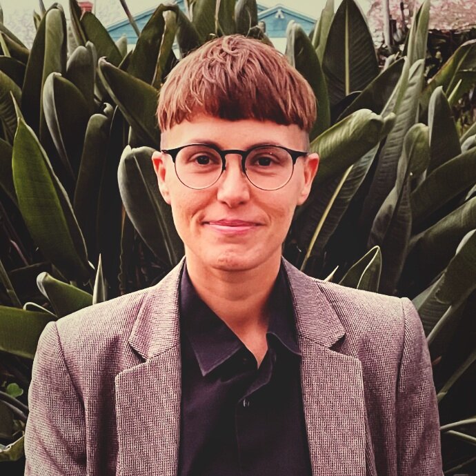 Una foto de AJ Schaerer, una persona trans, con una camisa abotonada y una chaqueta de traje, parado frente a una planta verde.