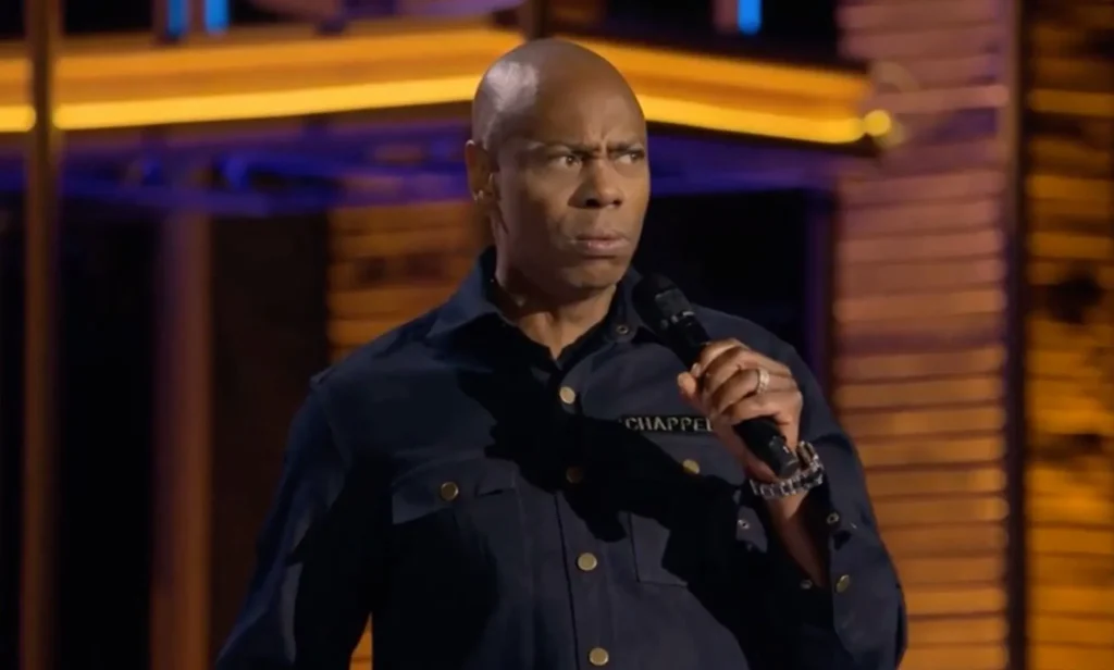 Dave Chappelle, who has faced backlash for making anti-trans jokes, wears a dark shirt as he speaks into a microphone in his new Netflix special The Dreamer