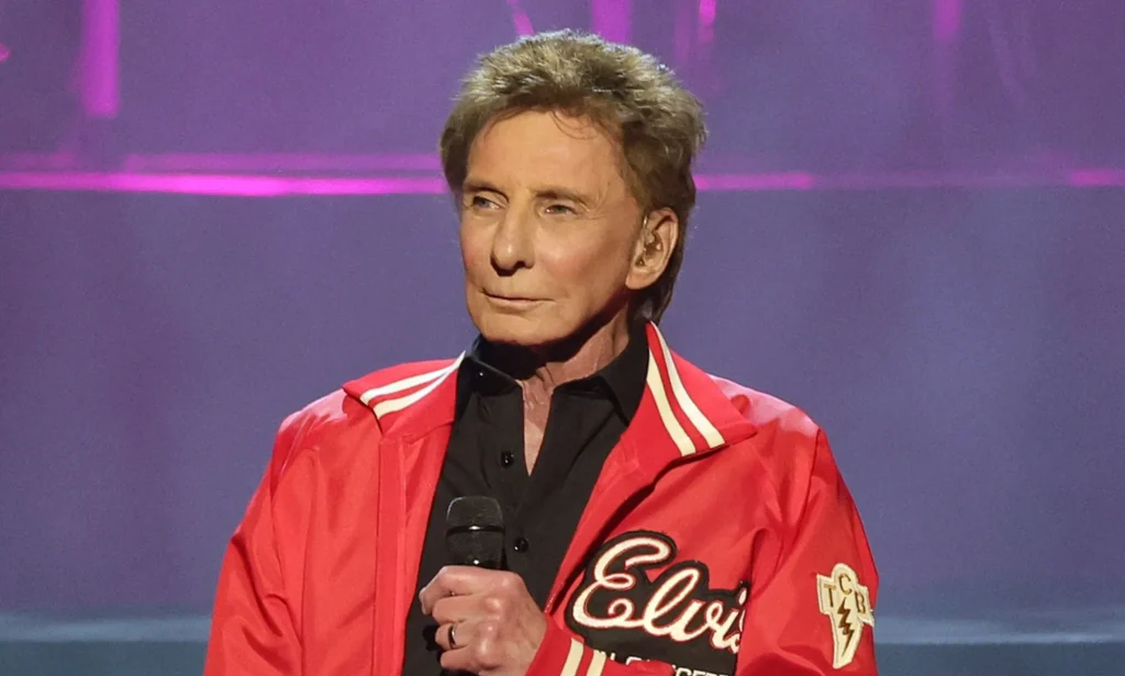 Singer Barry Manilow wears a black shirt and red jacket as he holds a microphone in front of his chest