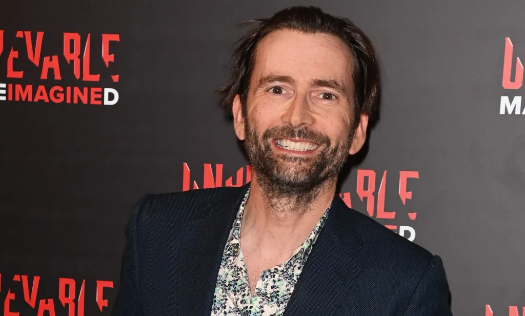 David Tennant, who is set to host the 2024 BAFTAs, smiles while wearing a black and white patterned shirt and dark jacket