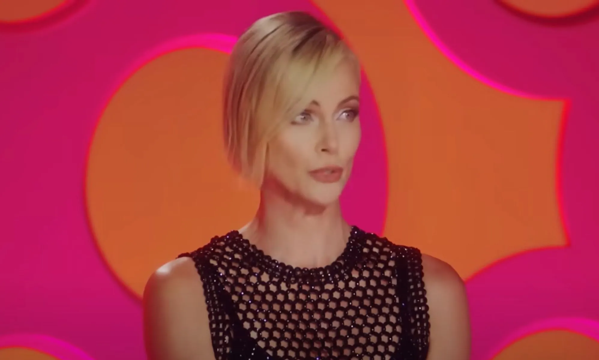 Charlize Theron wears a black outfit as she sits in front of a pink and orange background as a guest judge on Drag Race