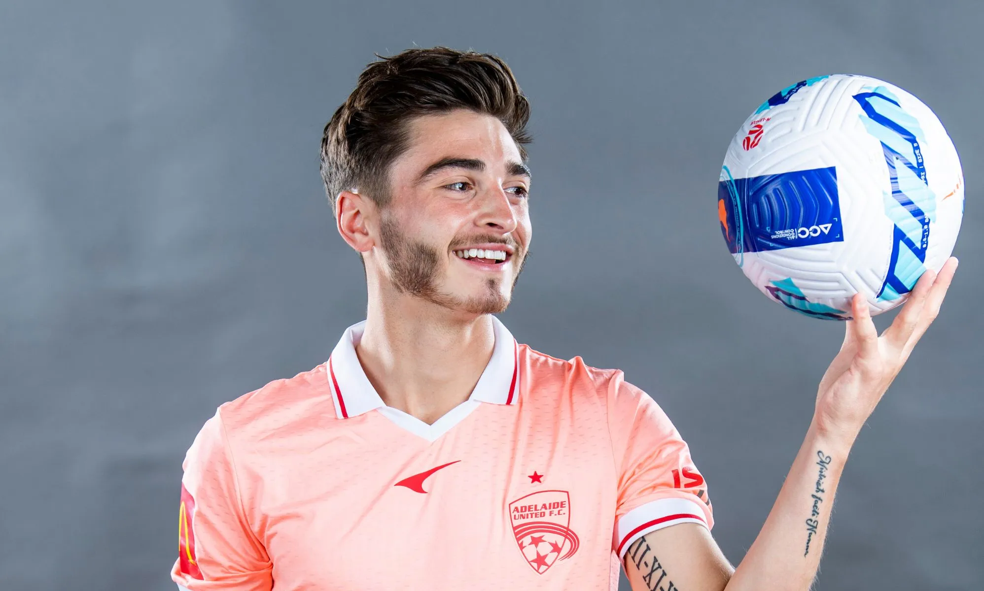 Josh Cavallo wears a pale pink Adelaide United football uniform as he smiles and holds up a blue, red and white football in one hand