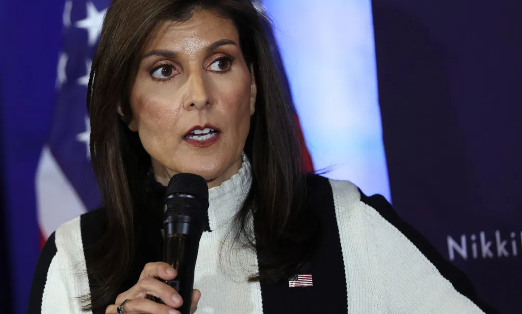 Nikki Haley viste una camisa blanca y un top negro con un pin de la bandera estadounidense mientras habla ante una multitud de seguidores fuera de cámara en el caucus de Iowa, donde intenta luchar contra Donald Trump por la nominación presidencial del partido.