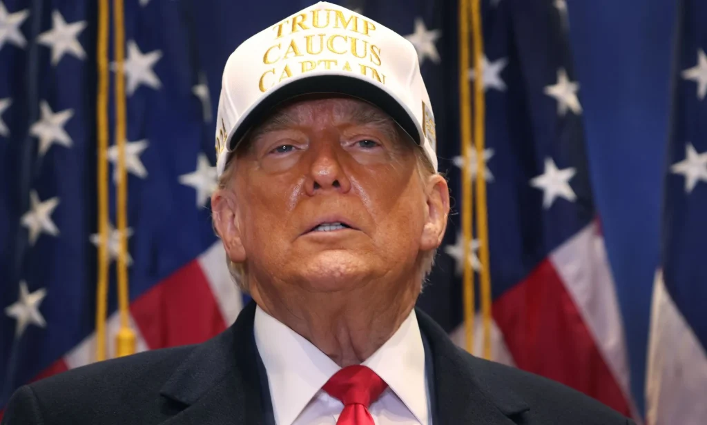 Donald Trump wears a suit and tie with a hat that reads