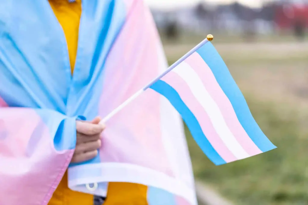 A trans person holding a flag with the trans flag draped over them.