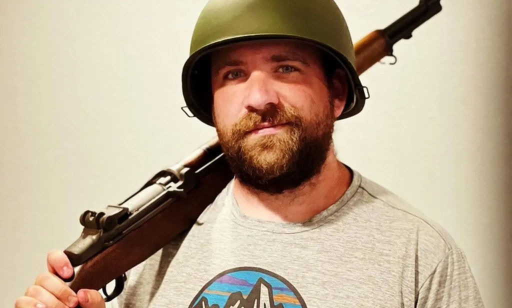 A man wearing a green army helmet holds a wooden rifle over his shoulder.
