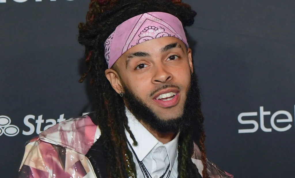 A man with dreadlocks and a chin strap beard wearing a pink bandana poses during a red carpet event.