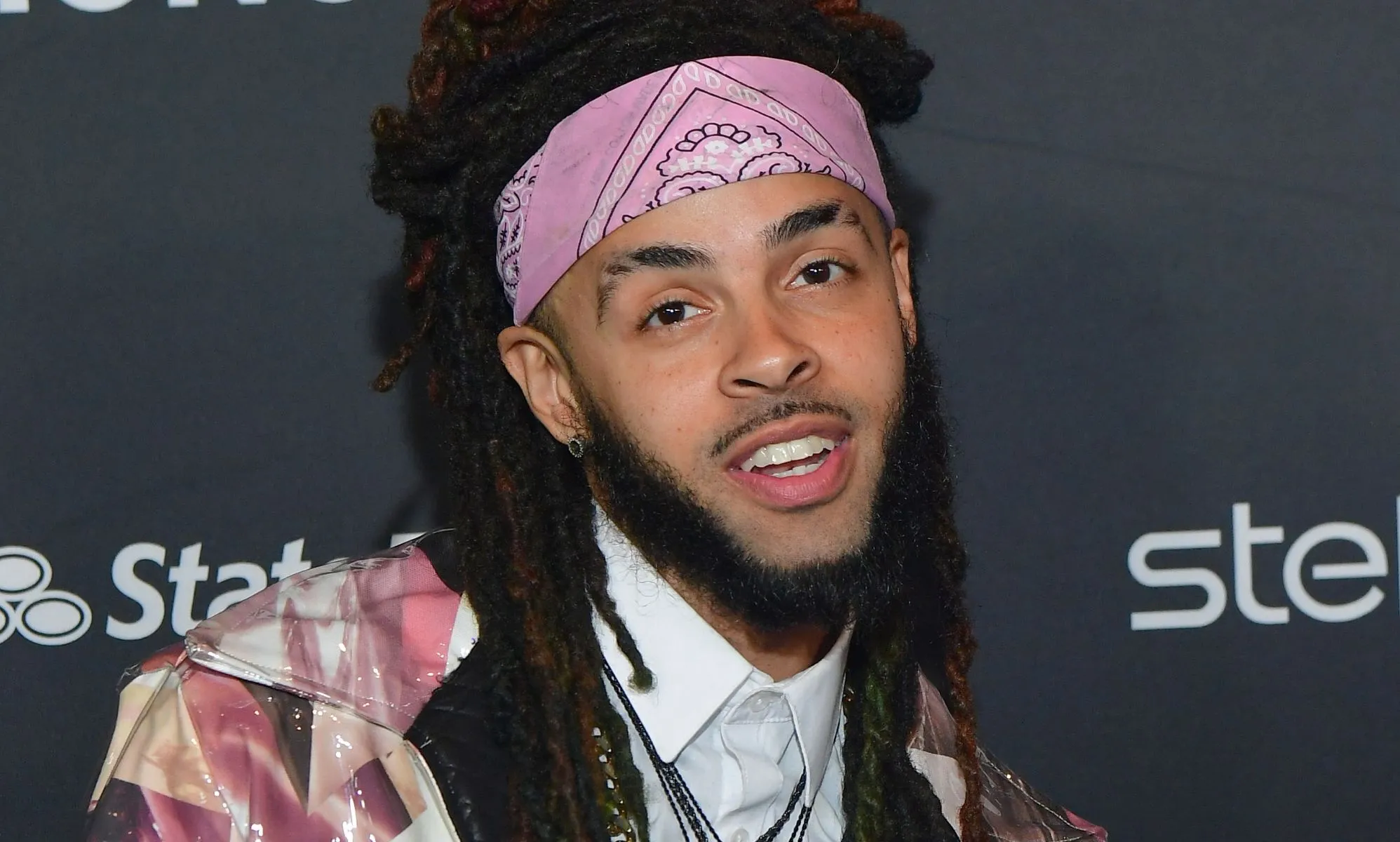 A man with dreadlocks and a chin strap beard wearing a pink bandana poses during a red carpet event.