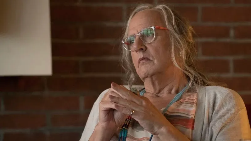 Jeffrey Tambor como el personaje trans Maura en Transparent.  Maura es una mujer mayor, lleva gafas, un cárdigan gris y un top de rayas rojas.
