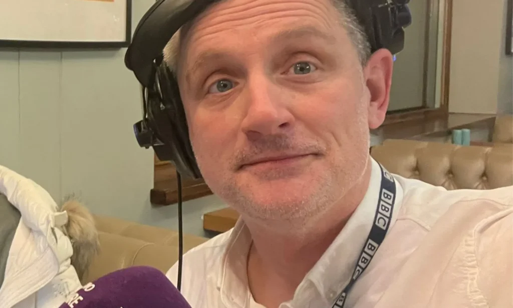 A man in a white button shirt wearing a pair of headphones on one ear smiles.